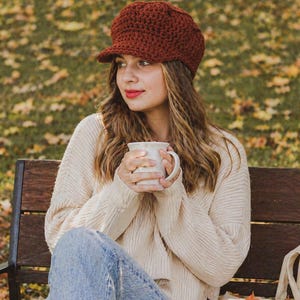 May include: A rust-colored crochet newsboy hat with a small brim. The hat is worn with a cream-colored sweater and blue jeans. A white mug is held in the hands. The background is a blurred view of fall foliage.
