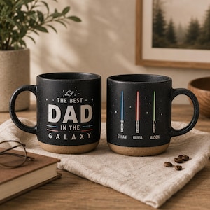 May include: Two black ceramic mugs with a speckled texture and a tan base. One mug features the text THE BEST DAD IN THE GALAXY with a small planet icon. The other mug displays three vertical lightsabers in blue, red, and green with the names ETHAN, OLIVIA, and MASON printed below them.