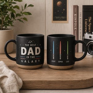 May include: Two black ceramic mugs with a tan base. The first mug features the text THE BEST DAD IN THE GALAXY with a small planet icon. The second mug displays three vertical lightsabers in blue, red, and green with the names HENRY, SOPHIA, and LUCA.