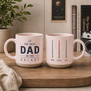 May include: Two light pink ceramic mugs with a speckled finish. One mug features the text THE BEST DAD IN THE GALAXY with a small planet icon. The other mug displays three vertical lightsabers in blue, red, and green with the names HENRY, SOPHIA, and LUCA.