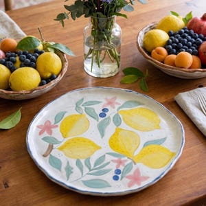 May include: A white ceramic serving platter with a hand-painted lemon design. The platter features yellow lemons, blue blueberries, pink flowers, and green leaves. The rim is outlined with a blue border. The platter is on a wooden table with bowls of fruit and a vase of flowers.