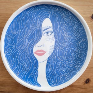 May include: A white ceramic plate featuring a blue and white illustration of a woman's face. The woman has long blue hair, pink lips, and intricate white designs on her face. The plate has a white rim and a glossy finish.