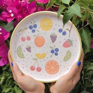 Könnte beinhalten: Ein runder weißer Teller mit gelbem Rand, der mit einem bunten Muster aus verschiedenen Früchten wie Orangen, Zitronen, Limetten, Kirschen, Erdbeeren, Blaubeeren und Brombeeren vor einem Hintergrund aus kleinen blauen Punkten verziert ist.