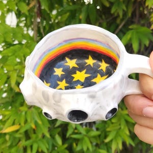 Op de afbeelding: Een witte keramische mok met een getextureerd, met kraters bedekt buitenoppervlak. De binnenkant heeft een zwarte basis beschilderd met gele sterren en een kleurrijke regenboogband rond de bovenrand, wat een levendig contrast vormt.