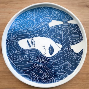 Può includere: Un piatto rotondo in ceramica con un'illustrazione bianca e blu scuro. Il design raffigura un volto con lunghi capelli fluenti che si fondono in onde oceaniche vorticose, accanto a un faro che emette un fascio di luce nel cielo scuro.