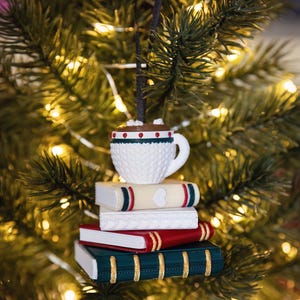 Può includere: Un ornamento sospeso che raffigura una tazza bianca testurizzata riempita di cioccolata calda e marshmallow, appoggiata su una pila di quattro libri. I libri sono color crema, bianco, rosso e verde scuro, con dettagli dorati sul dorso.