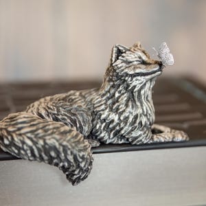 Peut inclure: Une figurine de renard en métal argenté en position de repos avec une finition en fourrure texturée. Un petit papillon délicat est posé sur le bout du nez du renard. La sculpture présente des détails complexes sur la fourrure et une surface lisse.