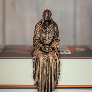 Puede incluir: Una figura de color bronce de un personaje encapuchado con una túnica larga y drapeada, sentado con las piernas colgando. El personaje tiene el rostro en sombra y sostiene un pequeño cuenco circular en las manos. La escultura tiene un acabado metálico.