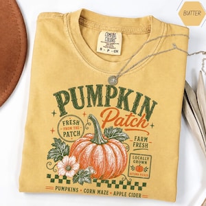 May include: A butter yellow t-shirt featuring a graphic of an orange pumpkin with green leaves and text that reads Pumpkin Patch, Fresh From The Patch, Farm Fresh, Locally Grown, Pumpkins, Corn Maze, and Apple Cider in green and orange ink.