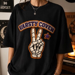 May include: A black short-sleeve t-shirt featuring a large chenille-style patch of a skeleton hand making a peace sign. Above the hand, the text VARSTY COVEN is embroidered in purple with an orange border. A small purple star is located to the right.