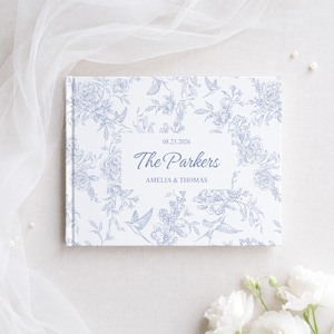Puede incluir: Un libro de tapa dura rectangular con una cubierta blanca que presenta un patrón floral y de aves en azul. Una etiqueta rectangular blanca central muestra el texto 08.23.2026, The Parkers y AMELIA & THOMAS en una fuente azul.