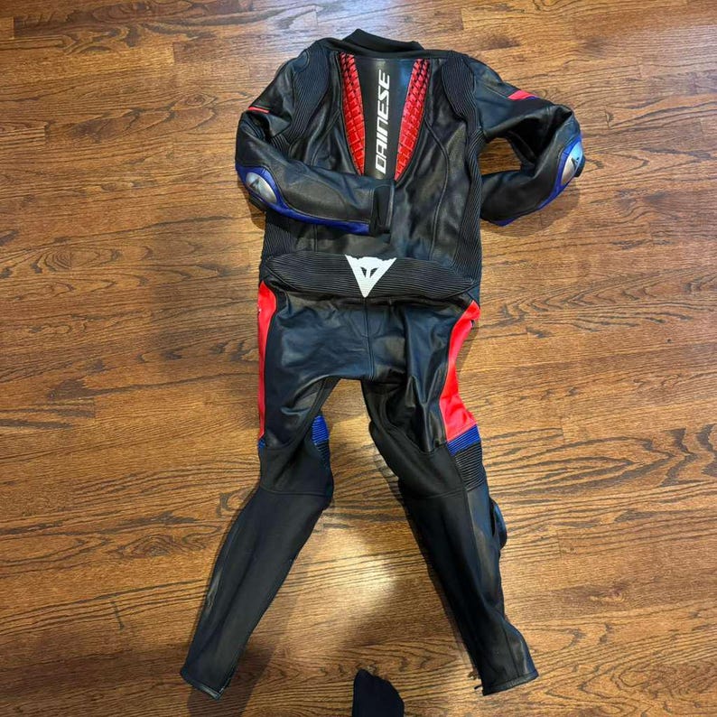 Puede incluir: Traje de carreras de moto negro y rojo con detalles azules y plateados. El traje tiene la marca "DAINESE" en la espalda y un logotipo triangular blanco en la parte delantera. Fabricado en cuero, incluye protecciones.