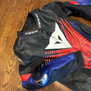 Puede incluir: Traje de carreras de moto negro y rojo con detalles azules y blancos. El traje presenta el logotipo "Dainese" en blanco en el cuello y el pecho. El traje tiene un dise&ntilde;o geom&eacute;trico en el pecho y los brazos.