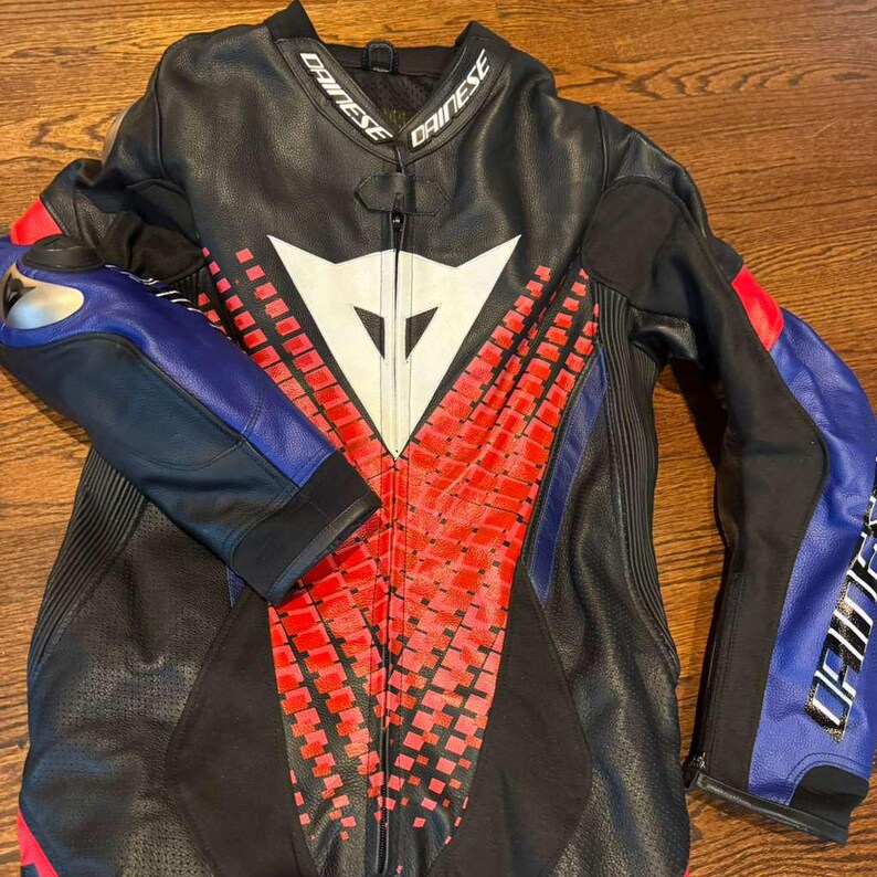 Puede incluir: Traje de carreras de moto negro y rojo con detalles azules en las mangas. El traje presenta un logotipo blanco en el pecho y el nombre de la marca "DAINESE" en el cuello. El traje es de cuero y tiene cierre de cremallera.