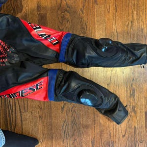 Puede incluir: Pantalones de moto negros y rojos con detalles azules. Los pantalones tienen protectores de rodilla y la palabra "DAINESE" es visible. Fabricados en cuero y otros materiales.
