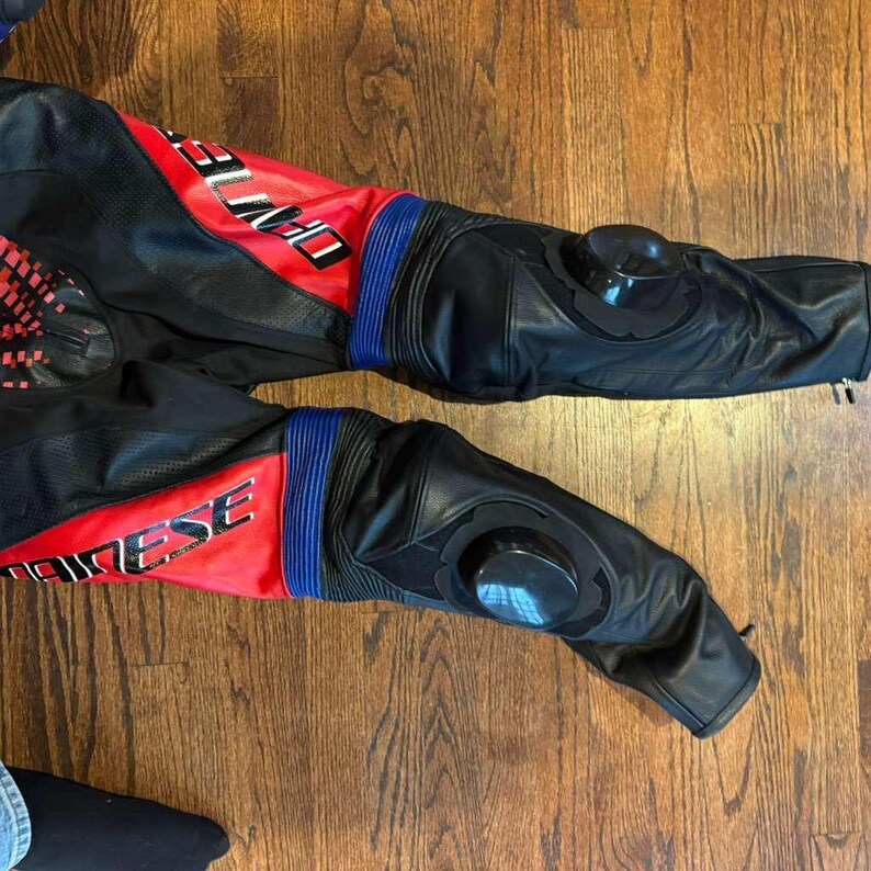 Puede incluir: Pantalones de moto negros y rojos con detalles azules. Los pantalones tienen protectores de rodilla y la palabra "DAINESE" es visible. Fabricados en cuero y otros materiales.