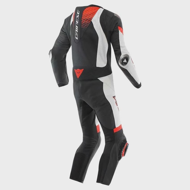 May include: A black, white and red motorcycle racing suit featuring the Dainese brand name on the upper back. The suit includes protective armour elements on the elbows and knees, with a perforated red panel on the upper back and a red logo on the waist.