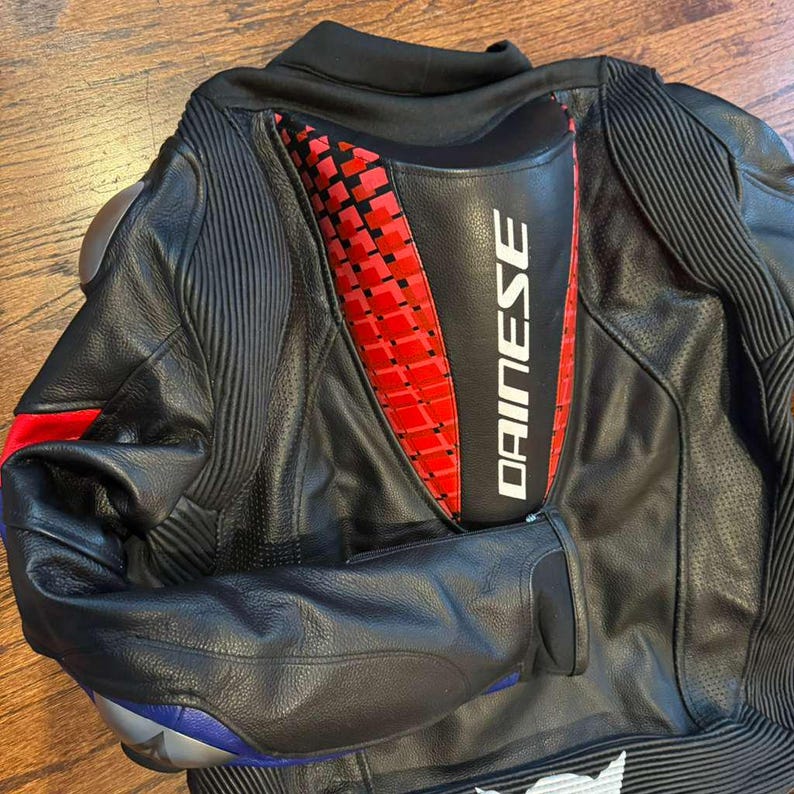 Puede incluir: Chaqueta de moto de cuero negro con detalles rojos y blancos. La espalda presenta un dise&ntilde;o geom&eacute;trico rojo y negro y el nombre de la marca "DAINESE" en blanco. La chaqueta tiene hombros acolchados y un detalle azul y plateado en la manga.