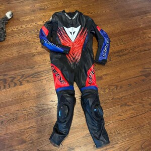 Puede incluir: Traje de carreras de moto negro y rojo con detalles azules. El traje presenta un logotipo blanco en el pecho y rodilleras. El nombre de la marca "Dainese" est&aacute; impreso en las mangas y las piernas.