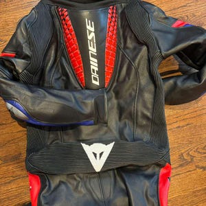 Puede incluir: Traje de moto de cuero negro con detalles rojos y blancos. El traje presenta el logotipo "DAINESE" en la espalda y un logotipo blanco en la parte inferior de la espalda. Detalles azules y rojos en las mangas y piernas.