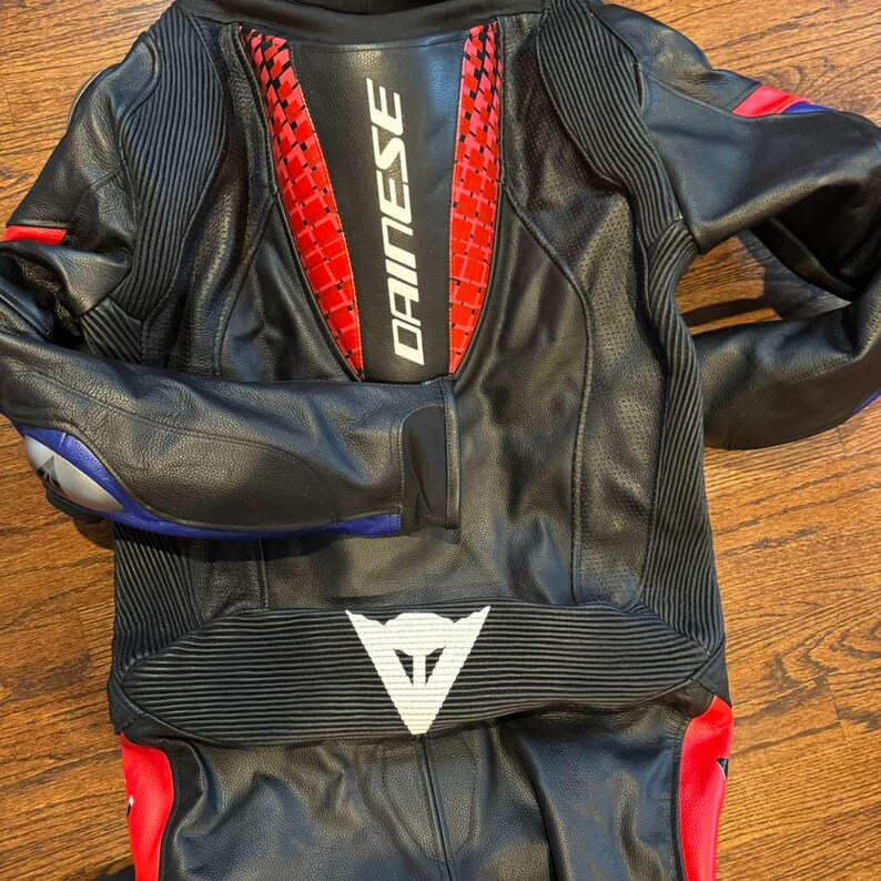 Puede incluir: Traje de moto de cuero negro con detalles rojos y blancos. El traje presenta el logotipo "DAINESE" en la espalda y un logotipo blanco en la parte inferior de la espalda. Detalles azules y rojos en las mangas y piernas.