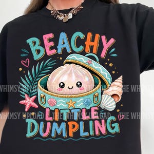 Può includere: T-shirt nero con una grafica colorata che recita "BEACHY LITTLE DUMPLING". Il design presenta un raviolo dei cartoni animati in una scatola di conchiglie, circondato da stelle marine, conchiglie e foglie tropicali. Il testo è in vari colori vivaci.