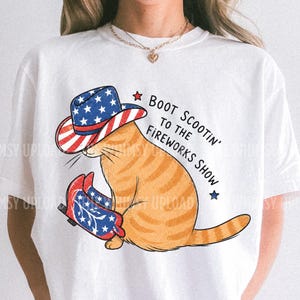 May include: White t-shirt featuring a cartoon orange tabby cat wearing a patriotic cowboy hat and boots. The hat and boots have a red, white, and blue design. Text on the shirt reads "Boot Scootin' to the Fireworks Show."