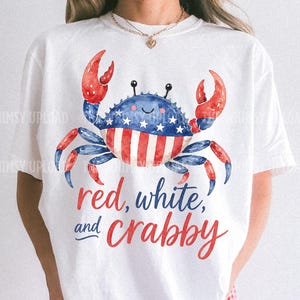 May include: White t-shirt featuring a watercolor crab design in red, white, and blue, with the text "red, white, and crabby." The crab has a blue body with white stars and red and white striped claws and belly. The shirt is paired with red and white checkered shorts.