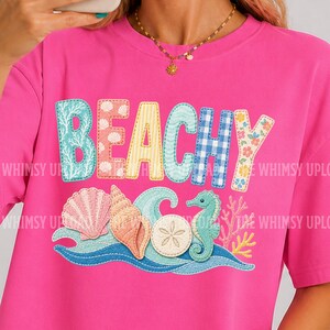 Op de afbeelding: Een felroze t-shirt met het woord "BEACHY" in kleurrijke, gestikte letters. Onder de tekst staan afbeeldingen van schelpen en zeepaardjes. Het shirt heeft korte mouwen en een ronde hals.
