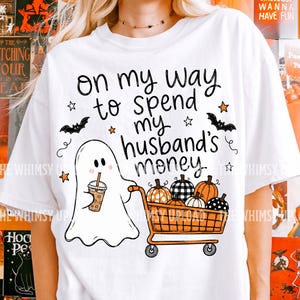 May include: A white t-shirt featuring a graphic of a ghost holding an iced coffee and pushing an orange shopping cart filled with various patterned pumpkins. The text reads, "on my way to spend my husband's money," accompanied by small black bats and orange stars.