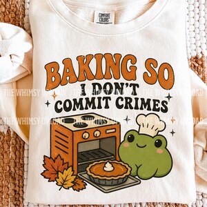 Könnte beinhalten: Ein cremefarbenes Sweatshirt mit Rundhalsausschnitt und einem Grafikdruck mit dem Text BAKING SO I DON'T COMMIT CRIMES. Das Design zeigt einen orangefarbenen Ofen, einen Kuchen, Herbstblätter und einen grünen Frosch mit einer weißen Kochmütze.