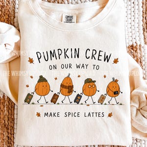May include: A cream-coloured crewneck sweatshirt featuring a graphic of four cartoon pumpkins walking with suitcases. The text reads PUMPKIN CREW ON OUR WAY TO MAKE SPICE LATTES. The pumpkins wear accessories like a beanie, scarf, glasses, and a camera.