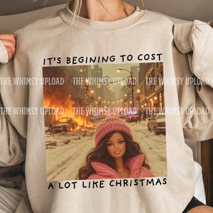 May include: A beige crewneck sweatshirt featuring a graphic print of a doll in a pink winter hat and jacket standing in a snowy city street with a fire in the background. The text reads IT'S BEGINING TO COST A LOT LIKE CHRISTMAS.