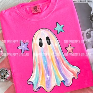 May include: A bright pink t-shirt featuring a central graphic of a pastel rainbow-colored ghost with black oval eyes. The ghost is surrounded by three small stars in light blue, purple, and peach. The shirt has a crew neckline and a Comfort Colors label.