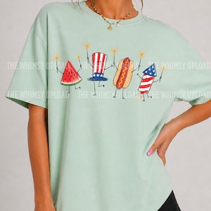 May include: A light green t-shirt featuring a whimsical illustration of a watermelon slice, Uncle Sam hat, hot dog, and firework, each holding a sparkler. The text "THE WHIMSY UPLOAD" is printed on the shirt.