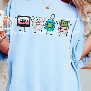 May include: A light blue t-shirt featuring a graphic print of four anthropomorphic 90s items: a black cassette tape, a rainbow-colored compact disc, a blue digital pet, and a grey handheld gaming console, all with smiling faces and thin limbs.