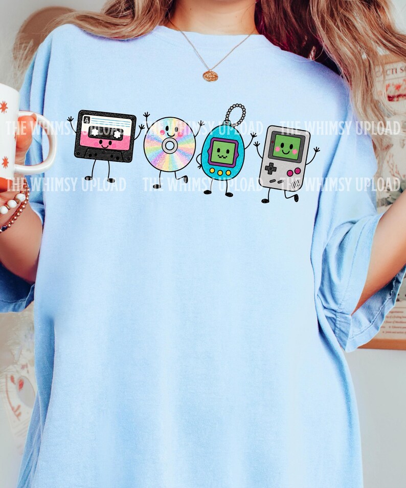 May include: A light blue t-shirt featuring a graphic print of four anthropomorphic 90s items: a black cassette tape, a rainbow-colored compact disc, a blue digital pet, and a grey handheld gaming console, all with smiling faces and thin limbs.