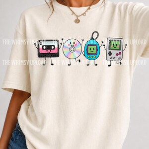 May include: A cream-colored short-sleeve t-shirt featuring a graphic print of four anthropomorphic retro electronics: a pink and black cassette tape, a rainbow-colored compact disc, a blue digital pet, and a grey handheld gaming console.