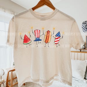 May include: A beige t-shirt featuring a whimsical illustration of a watermelon slice, a top hat, a hot dog, and a firework, each holding a sparkler. The design is in red, white, and blue, evoking a patriotic theme. The text "THE WHIMSY UPLOAD" is printed on the shirt.