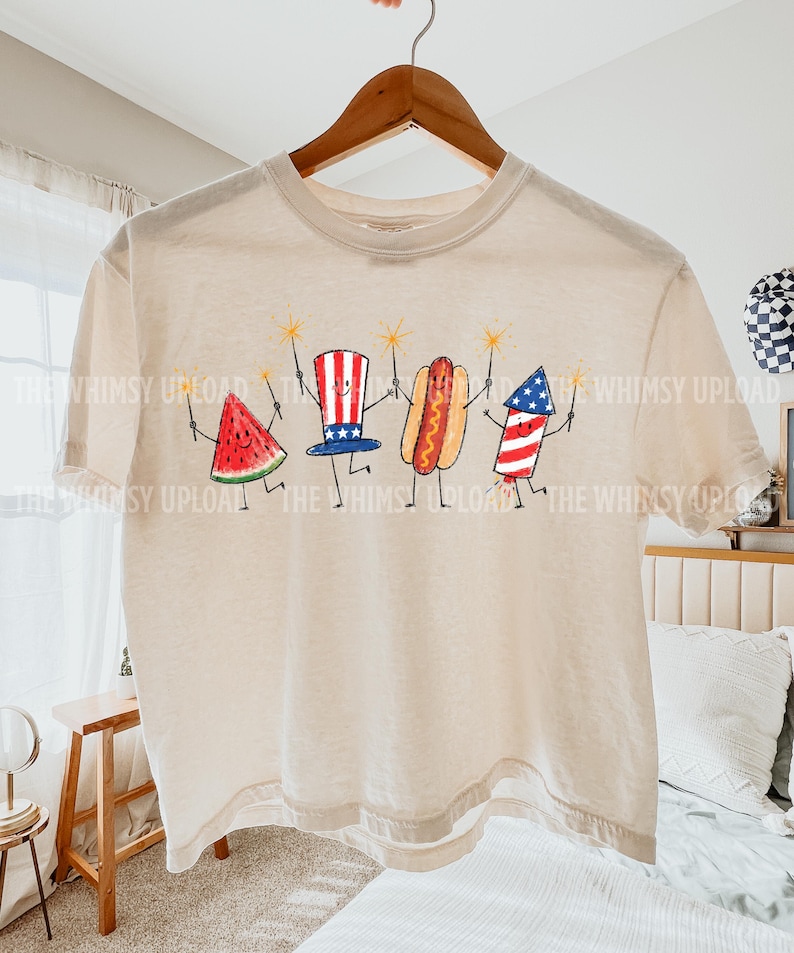 May include: A beige t-shirt featuring a whimsical illustration of a watermelon slice, a top hat, a hot dog, and a firework, each holding a sparkler. The design is in red, white, and blue, evoking a patriotic theme. The text "THE WHIMSY UPLOAD" is printed on the shirt.