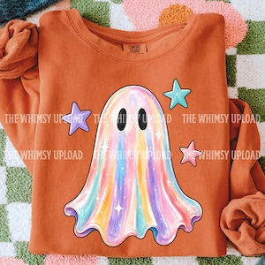 May include: An orange crewneck sweatshirt featuring a central graphic of a ghost with a multi-colored iridescent finish. The ghost is surrounded by three small stars in light blue, purple, and pink. The inner neck label reads Comfort Colors.