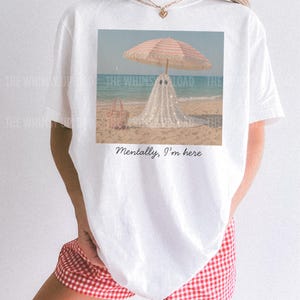 May include: White t-shirt with a beach scene print: a ghost under a striped parasol. A pink beach bag is visible. The text "Mentally, I'm here" is below the image. The shirt is worn with red and white checked shorts.