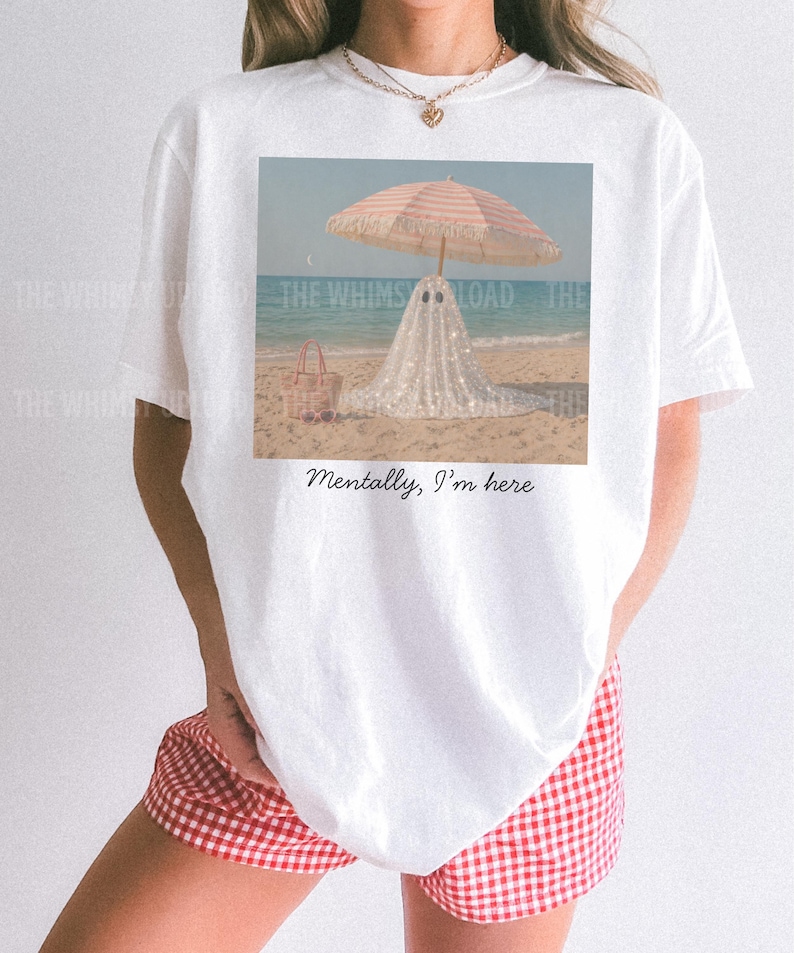 May include: White t-shirt with a beach scene print: a ghost under a striped parasol. A pink beach bag is visible. The text "Mentally, I'm here" is below the image. The shirt is worn with red and white checked shorts.