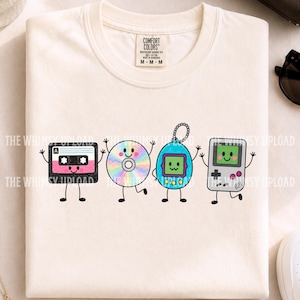 May include: A cream-colored t-shirt featuring a graphic print of four anthropomorphic retro items: a black cassette tape, a rainbow-colored compact disc, a blue digital pet, and a grey handheld gaming console, all with smiling faces and limbs.