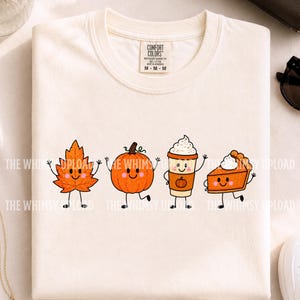 May include: A cream-colored t-shirt featuring a graphic of four cute, smiling autumn-themed characters: an orange maple leaf, a round pumpkin, a coffee cup with whipped cream, and a slice of pumpkin pie, all with thin black arms and legs.