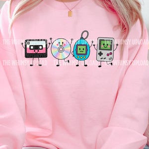 May include: A light pink crewneck sweatshirt featuring a graphic print of four anthropomorphic retro electronics: a black cassette tape, a rainbow-colored compact disc, a blue digital pet keychain, and a grey handheld video game console.