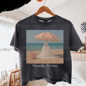 May include: Dark grey t-shirt with a beach scene print. A ghost in a sparkly white sheet stands under a striped parasol. A straw bag is on the sand. The text "Mentally, I'm here" is printed on the shirt.
