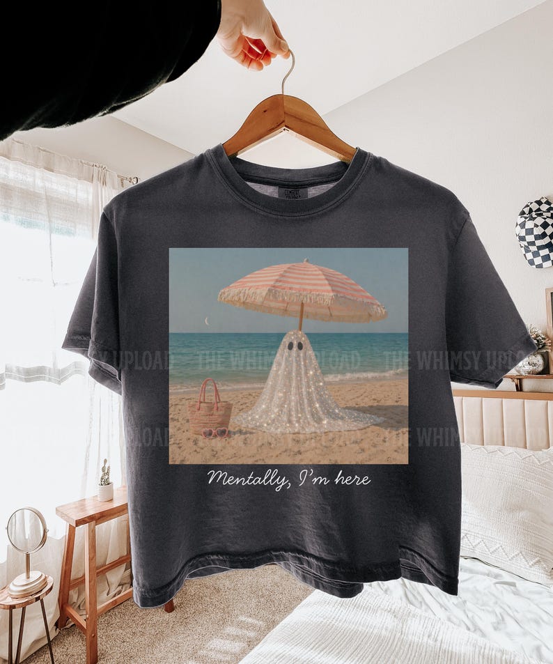 May include: Dark grey t-shirt with a beach scene print. A ghost in a sparkly white sheet stands under a striped parasol. A straw bag is on the sand. The text "Mentally, I'm here" is printed on the shirt.