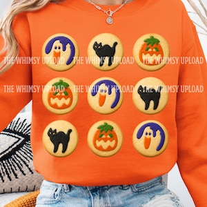May include: An orange crewneck sweatshirt featuring a repeating pattern of nine round cookies decorated with icing. The designs include a purple ghost, a black cat, and an orange jack-o'-lantern. The text THE WHIMSY UPLOAD is printed across the design.