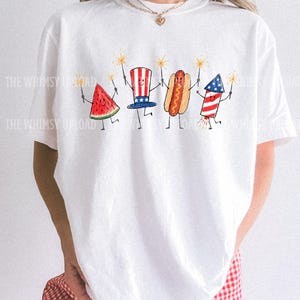 May include: White t-shirt featuring a whimsical illustration of a watermelon slice, a hot dog, a firework, and a patriotic hat, each holding sparklers. The design is in red, white, and blue, perfect for Independence Day celebrations.